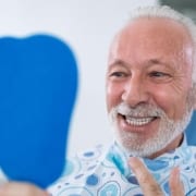 A senior man smiling while gazing into a handheld mirror to admire the results of his elderly dental care services